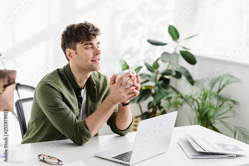 Canvas-taulu handsome businessman in green shirt sitting, holding cup near table with laptop