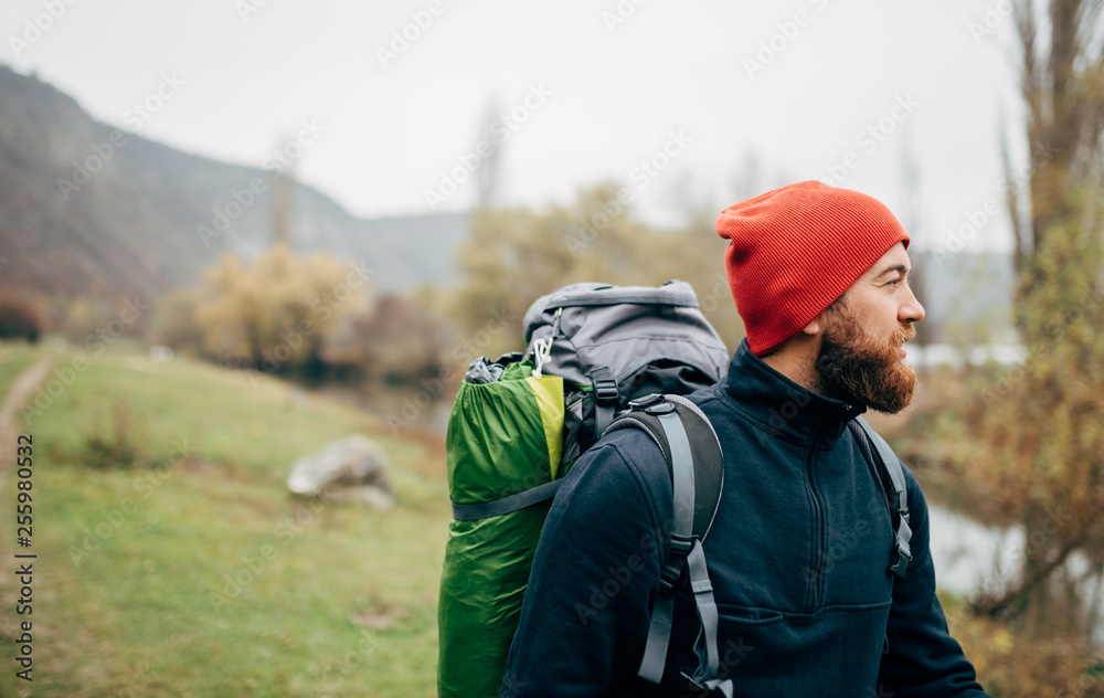 Outdoors horizontal portrait of young man hiking in mountains with travel backpack looking to a beautiful view. Traveler bearded man relaxing after trekking. Travel, people and lifestyle concept