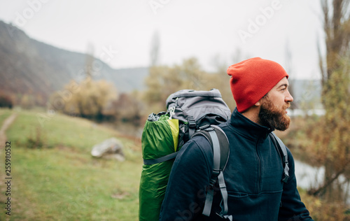 Outdoors horizontal portrait of young man hiking in mountains with travel backpack looking to a beautiful view. Traveler bearded man relaxing after trekking. Travel, people and lifestyle concept