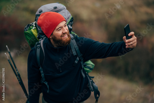 Happy young man smiling, taking selfie in mountains from his smart phone. Traveler bearded male wearing red hat take self portrait from mobile phone after hiking. Travel, lifestyle