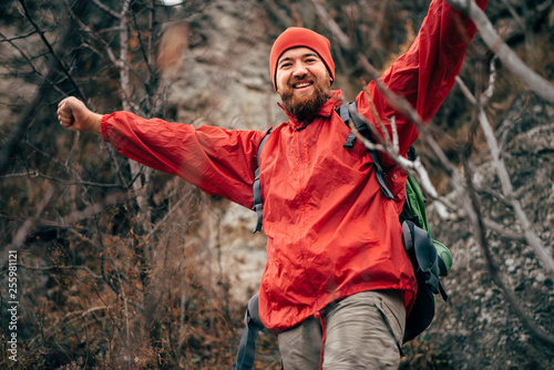 Portrait of happy young hiker man hiking in mountains wearing red clothes exploring new place. Traveler bearded man trekking and mountaineering during his journey. Travel, people, sport and lifestyle