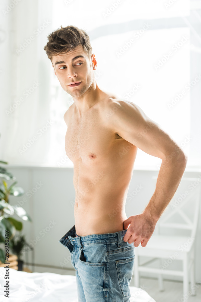 handsome man with bare torso standing near window and putting on blue jeans at home handsome man with bare torso standing near window and putting on blue jeans at home
