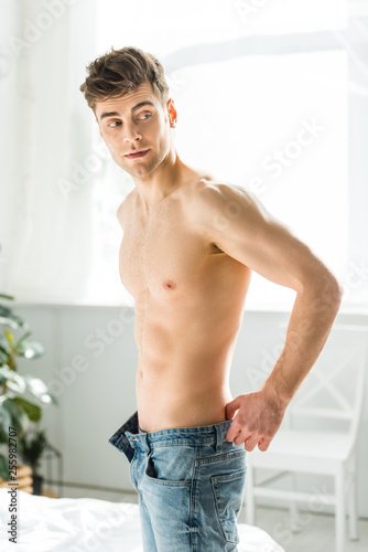 handsome man with bare torso standing near window and putting on blue jeans at home