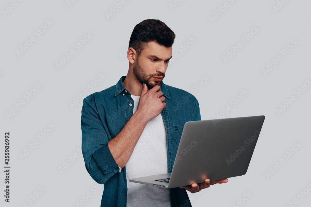 Considering next step. Thoughtful young man using laptop and keeping hand on chin while standing against grey background Considering next step. Thoughtful young man using laptop and keeping hand on chin while standing against grey background