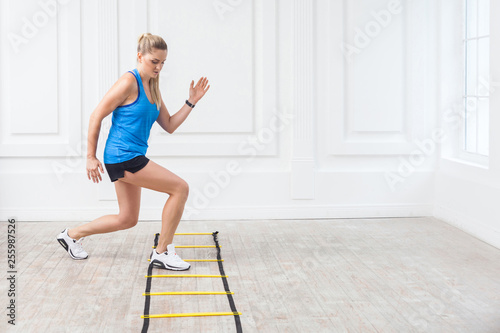 Full length of sporty beautiful young athletic blonde woman in black shorts and blue top are hard working and training on agility ladder drills in gym. Indoor, studio shot, workout and sport concept.