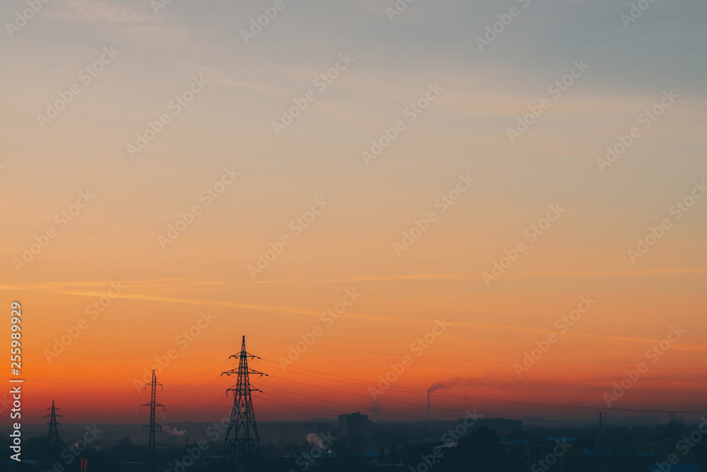 Power lines in city on dawn. Silhouettes of urban buildings among smog on sunrise. Cables of high voltage on warm orange yellow sky. Power industry at sunset. City power supply. Mist urban background.