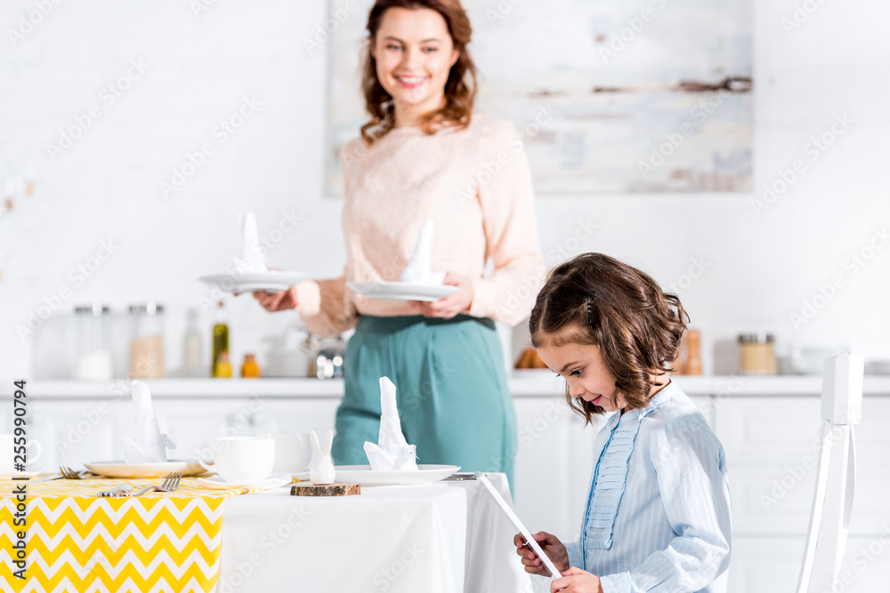 Child using digital tablet while mother serving table in kitchen Child using digital tablet while mother serving table in kitchen