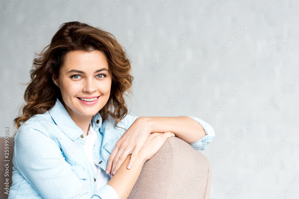 Happy brunette woman in shirt sitting on sofa and showing okay sign on grey Happy brunette woman in shirt sitting on sofa and showing okay sign on grey