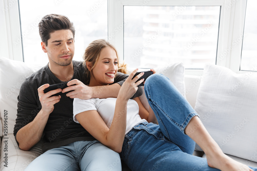 Portrait of european couple man and woman using mobile phones while lying on couch at home - obrazy, fototapety, plakaty 