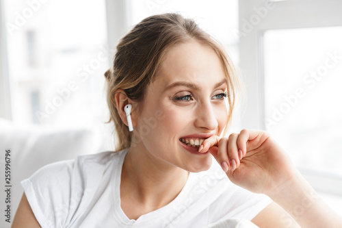 Portrait of blond woman wearing earpod smiling and sitting on couch in bright room at home
