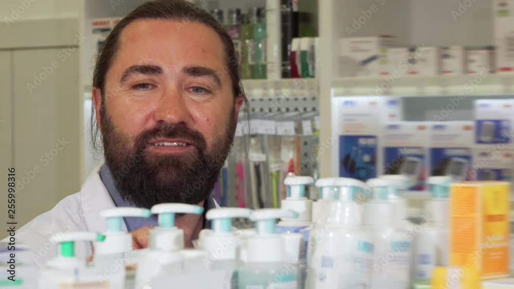 Bearded pharmacist smiling to the camera, while working at drugstore. Handsome mature bearded male chemist checking stock in an aisle, working at pharmacy. Job, doctor concept