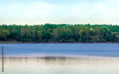 Quiet blue lake with slightly curled surface in front of a forest with dense tree population in front of a cloudy sky