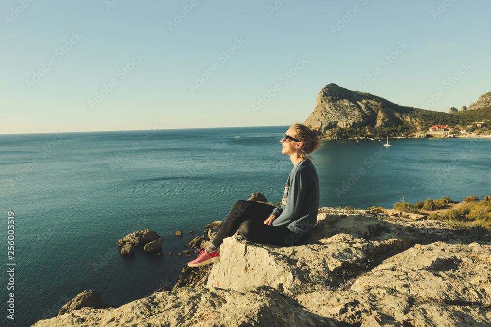 profile portrait of young happy girl with hair bun and sunglasses is sitting on the rocky precipice, edge of mountain, blue sea below