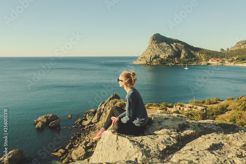 profile portrait of young girl with hair bun and sunglasses is sitting on the rocky precipice, edge of mountain, blue sea below