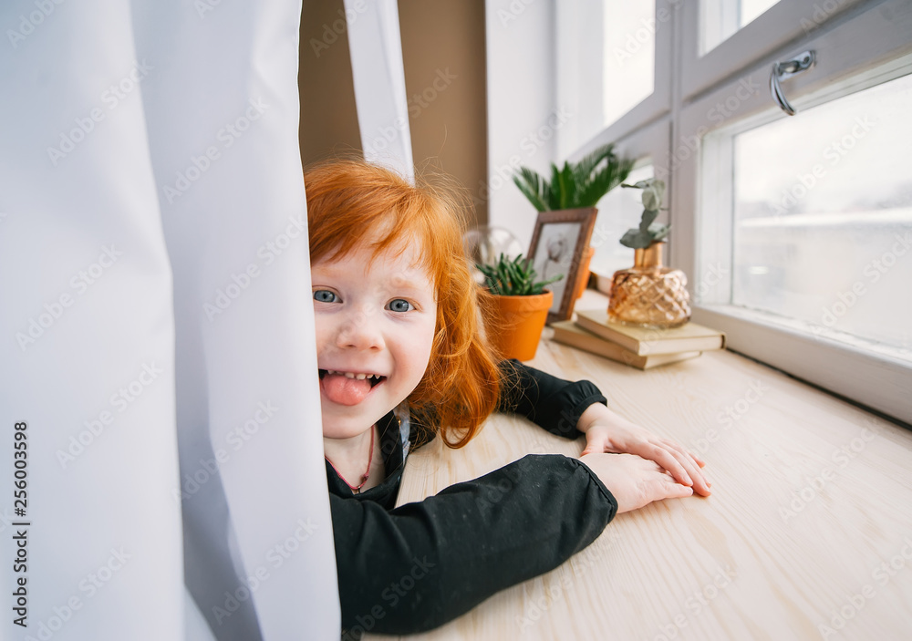 Pretty redhead litlle girl sitting at table near window and she puts out the tongue Pretty redhead litlle girl sitting at table near window and she puts out the tongue