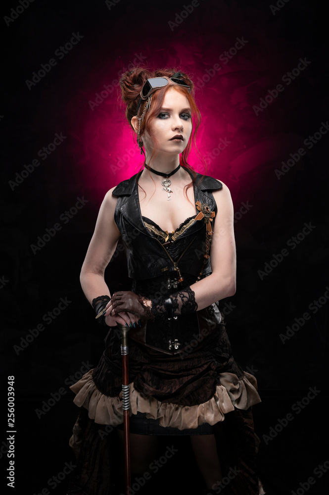 A beautiful red-haired cosplayer girl wearing a Victorian-style steampunk costume with large breasts in a deep neckline and glasses holding a cane on a black and purple background