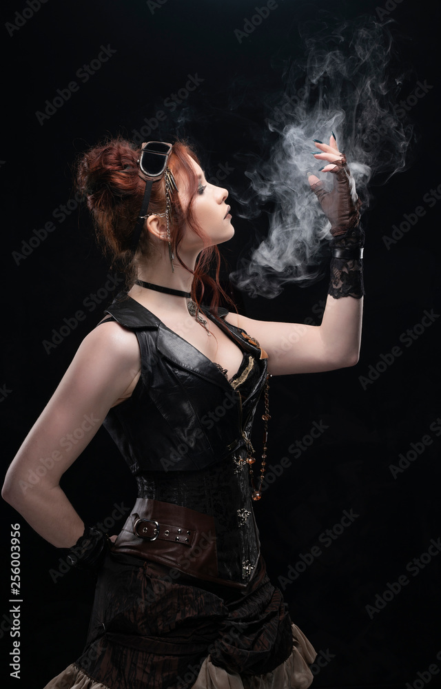 A beautiful redhead cosplayer girl wearing a Victorian-style steampunk costume with big breasts in a deep neckline holds her hand in a puff of smoke on a black background