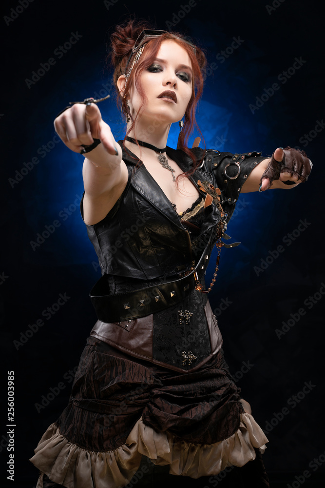 Fototapeta premium Beautiful redhead cosplayer girl wearing a Victorian-style steampunk costume with a big breast in a deep neckline points with both hands on a black and blue background.