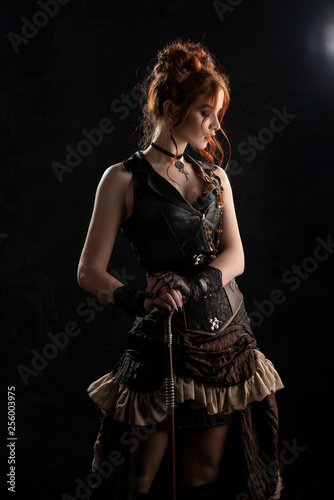 A beautiful red-haired cosplayer girl wearing a Victorian-style steampunk costume with a big breast in a deep neckline stands thoughtfully, leaning on a cane, on a dark background.