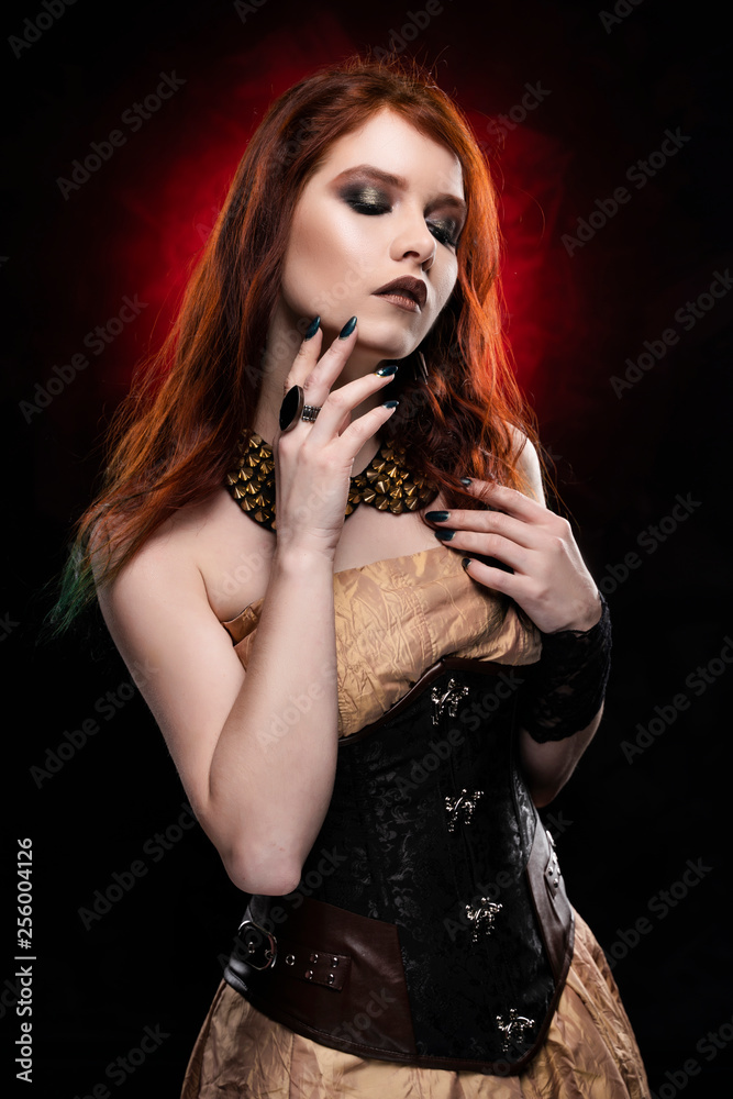 A beautiful thoughtful redhead cosplay girl wearing a Victorian style steampunk dress and corset. Portrait. Black and red background. foto de Stock Adobe Stock