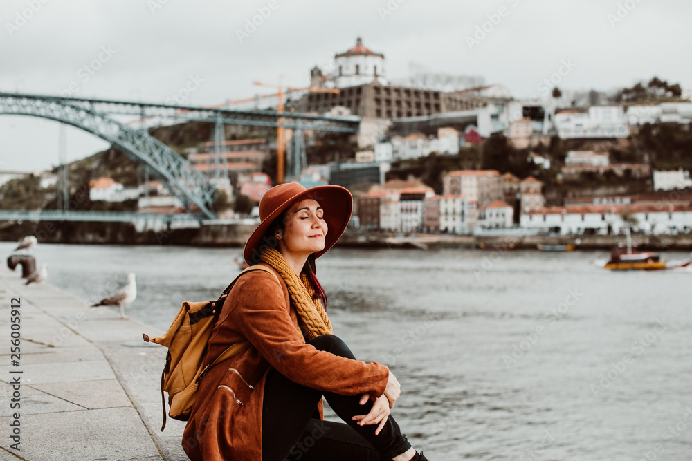 .Young and pretty woman sightseeing in the city of Porto, Portugal. Sitting on the river side, enjoying a beautiful landscape on a windy spring day. Lifestyle.
