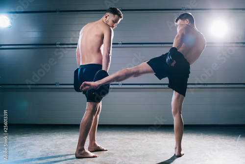 back view of strong muscular barefoot mma fighter practicing low kick with another sportsman