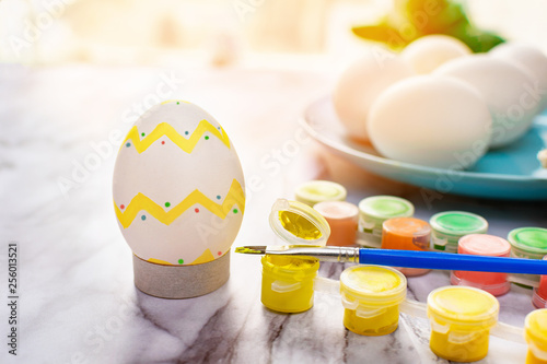 Easter concept - painted egg and colorful paints with a brush for the traditional painting of eggs, and a blue plate with unpainted eggs on a marble table on a sunny morning photo