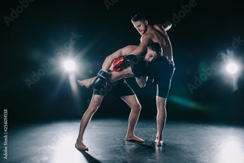 barefoot strong muscular mma fighter in boxing gloves clinching another while sportsman kicking him photo