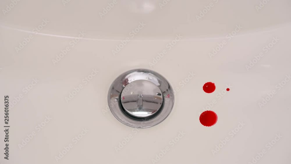 A close-up of blood drops falling into a white bathroom sink. Spots are washed by the stream of water A close-up of blood drops falling into a white bathroom sink. Spots are washed by the stream of water