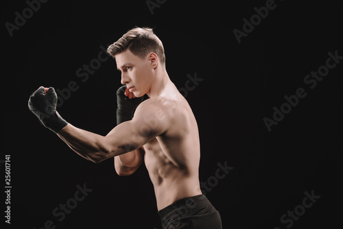 side view of athletic muscular sportsman in bandages doing punch isolated on black