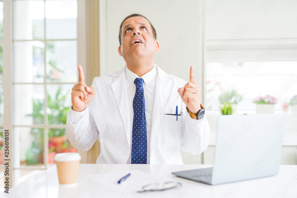Middle age doctor man wearing white medical coat working with laptop at the clinic amazed and surprised looking up and pointing with fingers and raised arms. Middle age doctor man wearing white medical coat working with laptop at the clinic amazed and surprised looking up and pointing with fingers and raised arms.