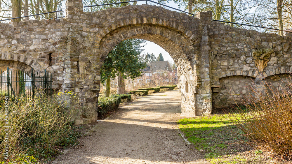 Stone bridge with an arch with a dirt pedestrian path in the Proosdij Park with small bushes, trees and houses in the background, sunny winter day in Meerssen, South Limburg, Netherlands