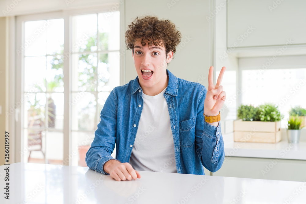 Young handsome man wearing casual denim jacket at home smiling with happy face winking at the camera doing victory sign. Number two. Young handsome man wearing casual denim jacket at home smiling with happy face winking at the camera doing victory sign. Number two.