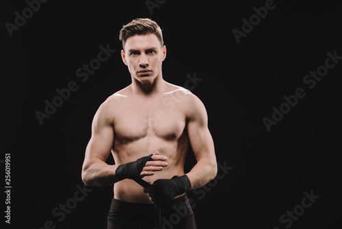 strong shirtless muscular mma fighter fixing bandages and looking at camera isolated on black