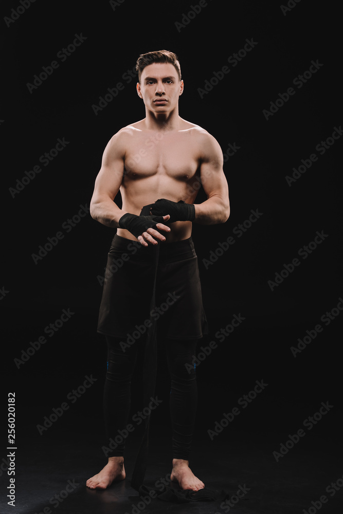 strong shirtless and barefoot muscular mma fighter fixing bandages and looking at camera on black