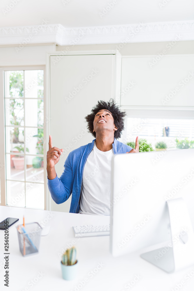 African American man working using computer amazed and surprised looking up and pointing with fingers and raised arms.