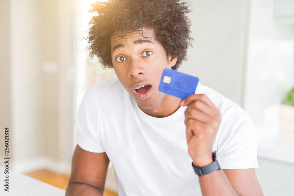 African American man holding credit card scared in shock with a surprise face, afraid and excited with fear expression African American man holding credit card scared in shock with a surprise face, afraid and excited with fear expression