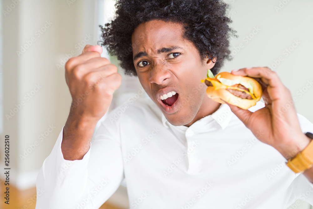 African American hungry man eating hamburger for lunch annoyed and frustrated shouting with anger, crazy and yelling with raised hand, anger concept African American hungry man eating hamburger for lunch annoyed and frustrated shouting with anger, crazy and yelling with raised hand, anger concept