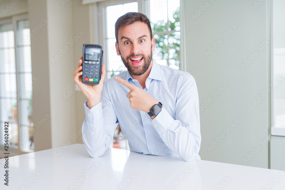 Handsome young man holding point of sale terminal very happy pointing with hand and finger