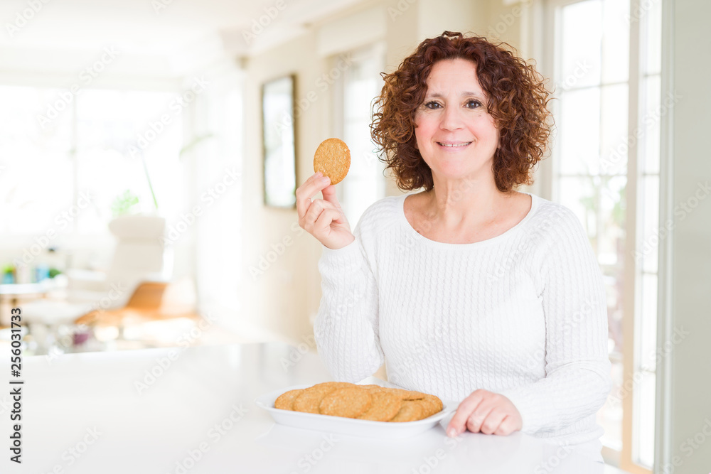 Senior woman eating healthy whole grain biscuit at home with a happy face standing and smiling with a confident smile showing teeth Senior woman eating healthy whole grain biscuit at home with a happy face standing and smiling with a confident smile showing teeth