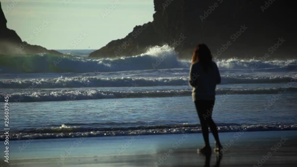 A woman bending over and picking up a shell from the beach as blue ocean waves roll towards the shoreline of the Oregon coast with large rocks in the background.