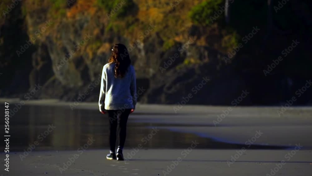 A young brunette woman in a grey sweater and black pants walking towards the calm water along the beach on the coast of Oregon with trees in the background in Autumn. Slow motion. A young brunette woman in a grey sweater and black pants walking towards the calm water along the beach on the coast of Oregon with trees in the background in Autumn. Slow motion.