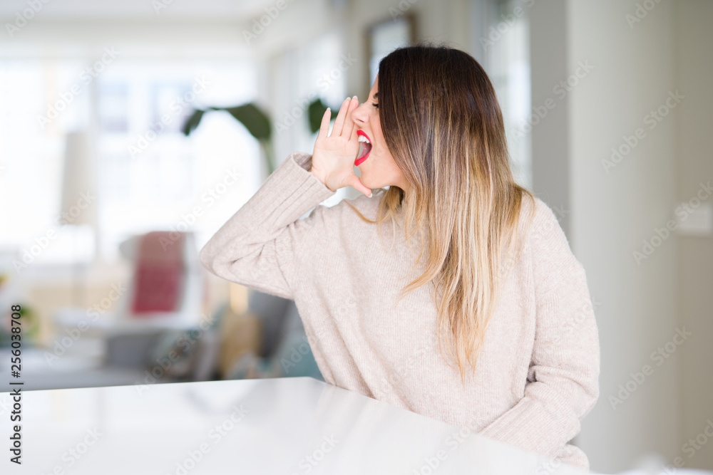 Young beautiful woman wearing winter sweater at home shouting and screaming loud to side with hand on mouth. Communication concept. Young beautiful woman wearing winter sweater at home shouting and screaming loud to side with hand on mouth. Communication concept.