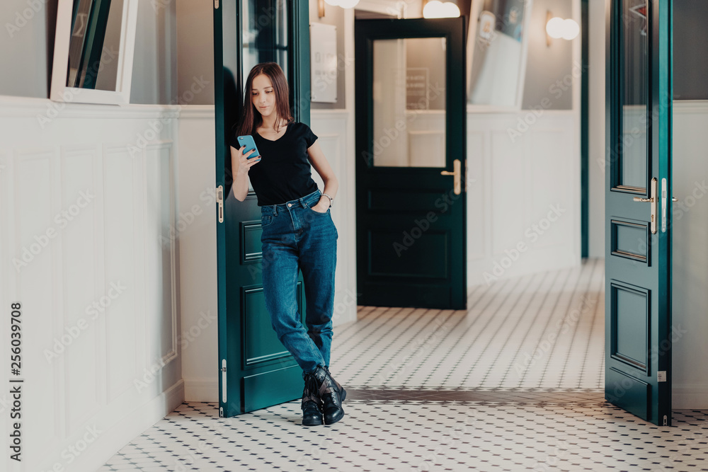 Full length shot of serious fashionable woman concentrated into cell phone, wears black t shirt, jeans and boots, reads information in internet, stands indoor near doors connected to wireless internet