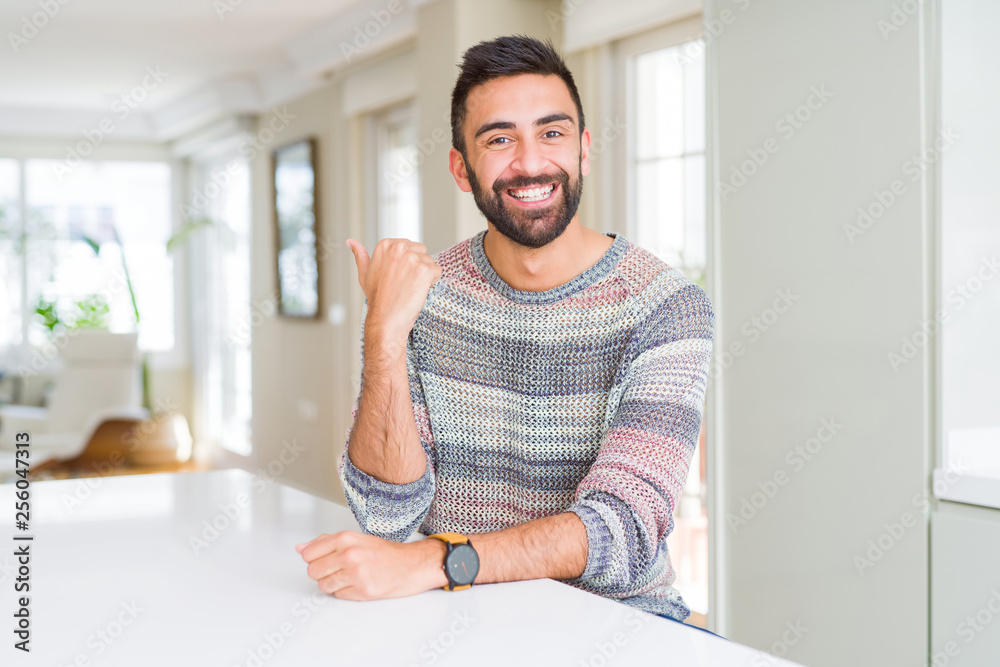 Handsome hispanic man wearing casual sweater at home smiling with happy face looking and pointing to the side with thumb up.