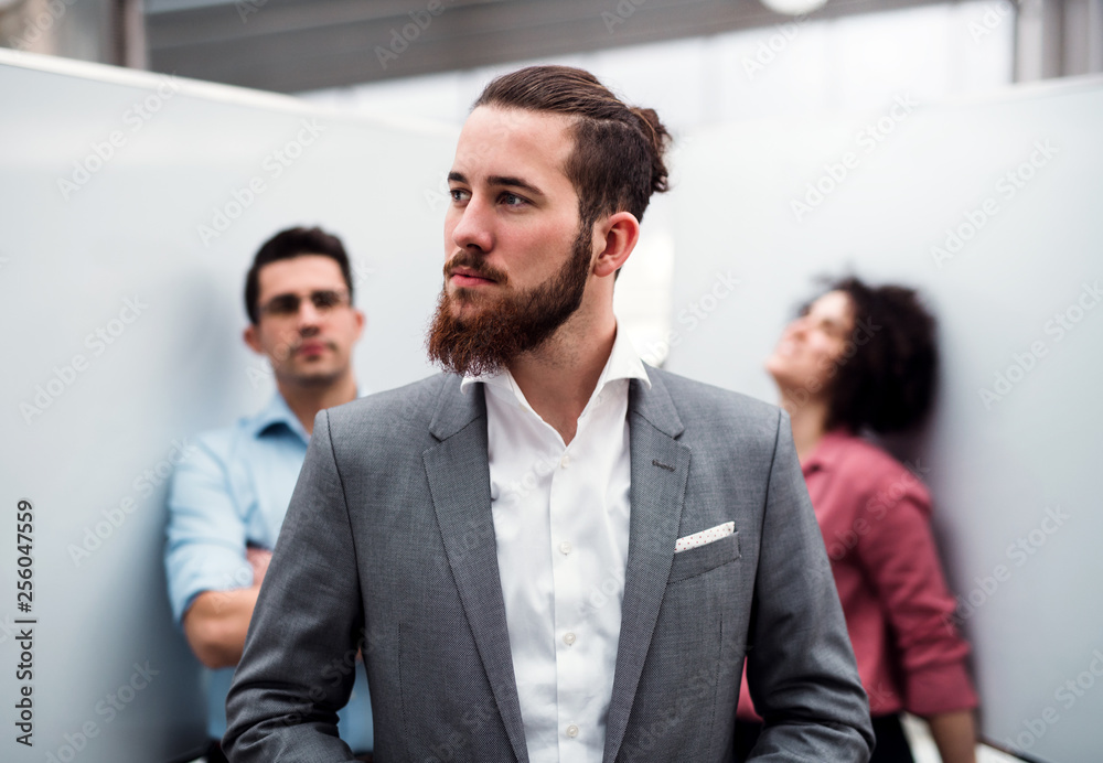 A young businessman in suit standing in office, colleagues in the background. A young businessman in suit standing in office, colleagues in the background.