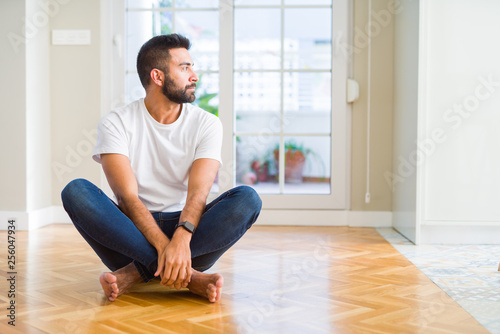 Handsome hispanic man wearing casual t-shirt sitting on the floor at home looking to side, relax profile pose with natural face with confident smile.