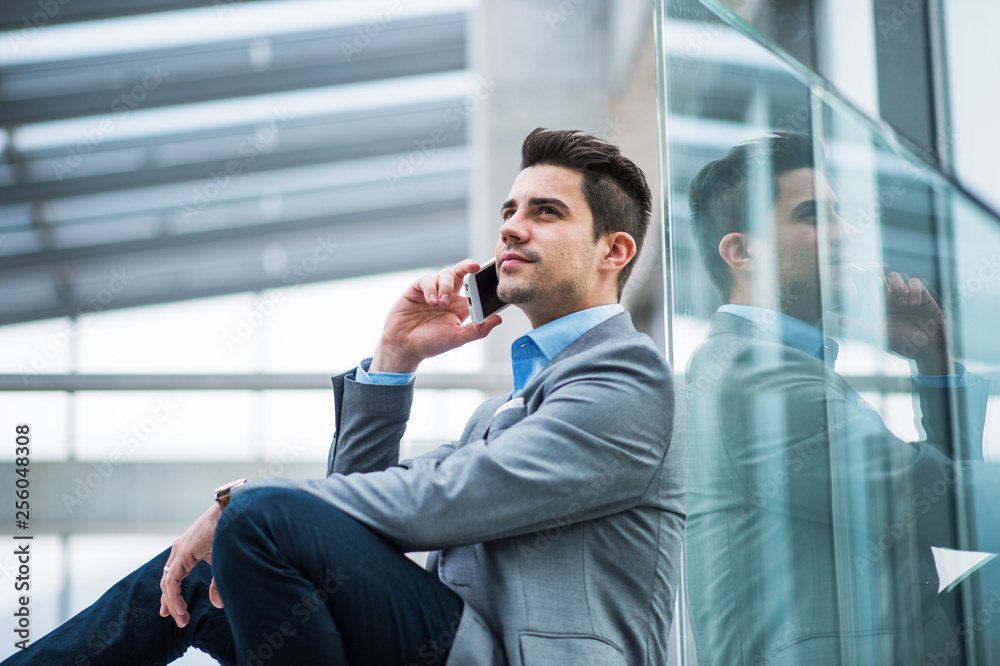 A portrait of young businessman with smartphone sitting in corridor outside office. A portrait of young businessman with smartphone sitting in corridor outside office.