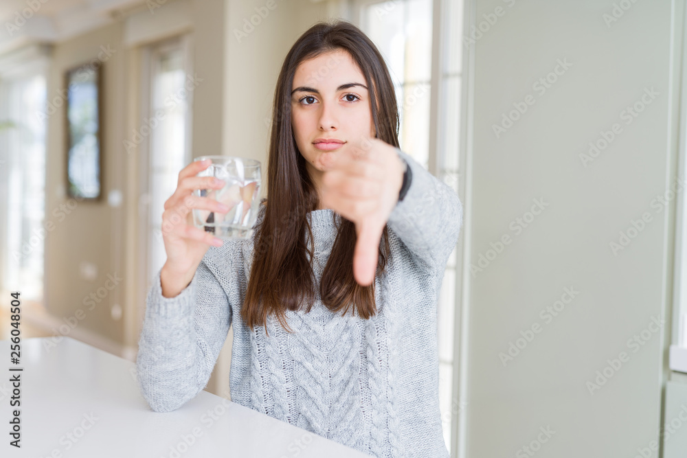 Beautiful young woman drinking a fresh glass of water with angry face, negative sign showing dislike with thumbs down, rejection concept Beautiful young woman drinking a fresh glass of water with angry face, negative sign showing dislike with thumbs down, rejection concept