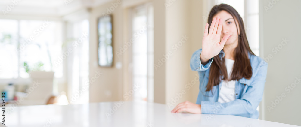 Wide angle picture of beautiful young woman sitting on white table at home doing stop sing with palm of the hand. Warning expression with negative and serious gesture on the face. Wide angle picture of beautiful young woman sitting on white table at home doing stop sing with palm of the hand. Warning expression with negative and serious gesture on the face.